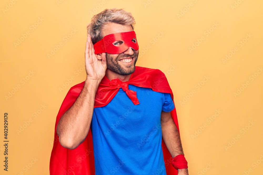 Handsome blond man wearing super hero costume with mask and cape over yellow background smiling with hand over ear listening and hearing to rumor or gossip. Deafness concept. Handsome blond man wearing super hero costume with mask and cape over yellow background smiling with hand over ear listening and hearing to rumor or gossip. Deafness concept.