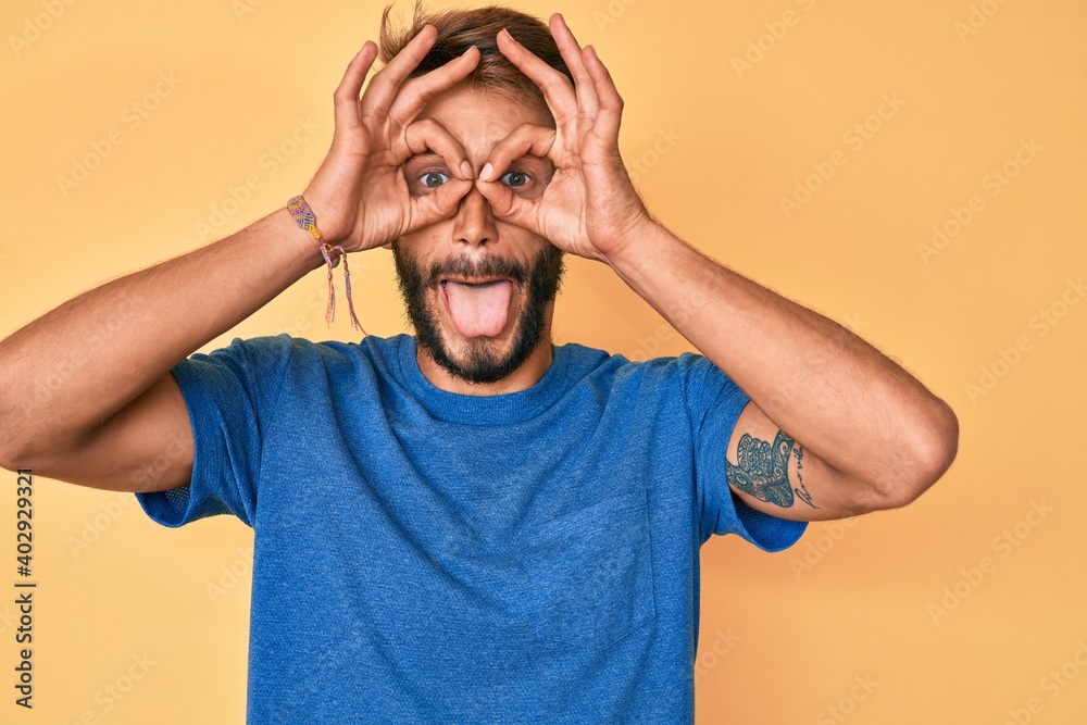 Handsome caucasian man with beard wearing casual clothes doing ok gesture like binoculars sticking tongue out, eyes looking through fingers. crazy expression. Handsome caucasian man with beard wearing casual clothes doing ok gesture like binoculars sticking tongue out, eyes looking through fingers. crazy expression.