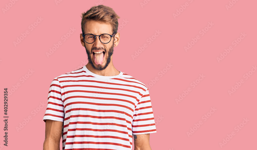 Handsome blond man with beard wearing casual clothes and glasses sticking tongue out happy with funny expression. emotion concept. Handsome blond man with beard wearing casual clothes and glasses sticking tongue out happy with funny expression. emotion concept.
