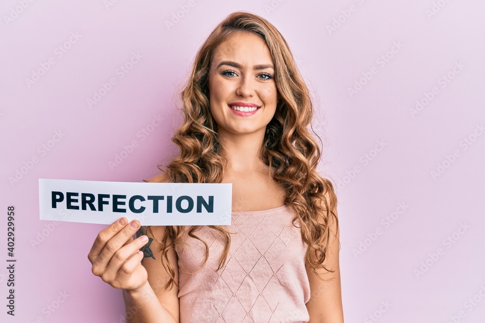 Young blonde girl holding paper with perfection message looking positive and happy standing and smiling with a confident smile showing teeth Young blonde girl holding paper with perfection message looking positive and happy standing and smiling with a confident smile showing teeth