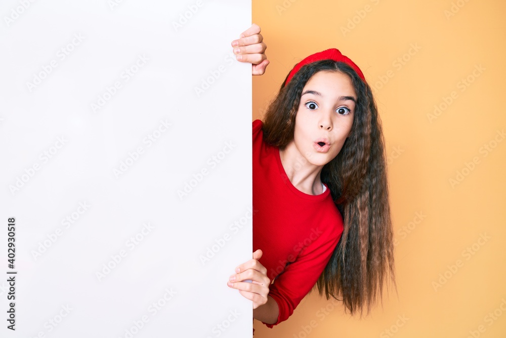 Cute hispanic child girl holding blank empty banner scared and amazed with open mouth for surprise, disbelief face