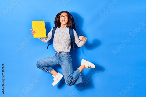 Young beautiful chinese student girl wearing glasses and backpack smiling happy. Jumping with smile on face holding book over isolated blue background
