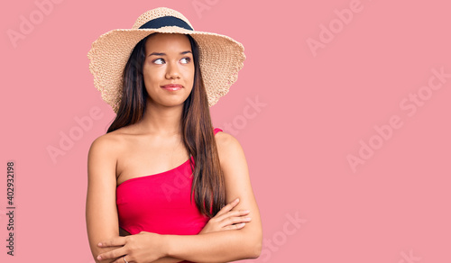 Young beautiful latin girl wearing swimwear and summer hat smiling looking to the side and staring away thinking.