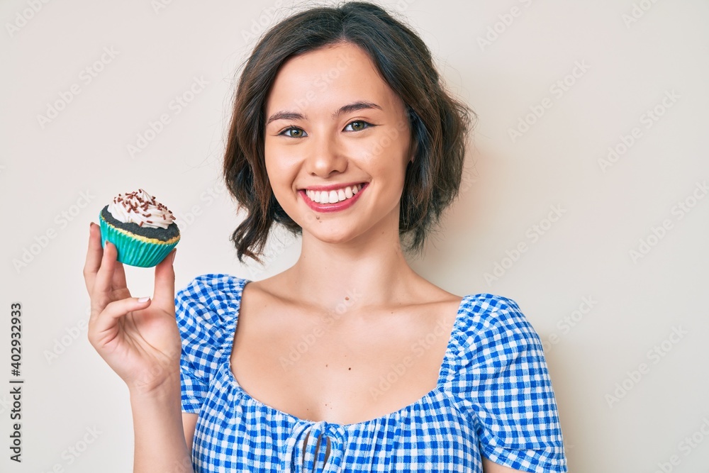 Young beautiful woman holding cupcake looking positive and happy standing and smiling with a confident smile showing teeth Young beautiful woman holding cupcake looking positive and happy standing and smiling with a confident smile showing teeth