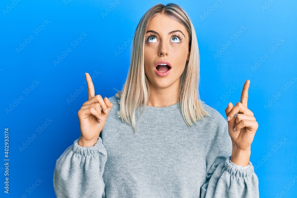 Beautiful blonde woman wearing casual clothes amazed and surprised looking up and pointing with fingers and raised arms. Beautiful blonde woman wearing casual clothes amazed and surprised looking up and pointing with fingers and raised arms.
