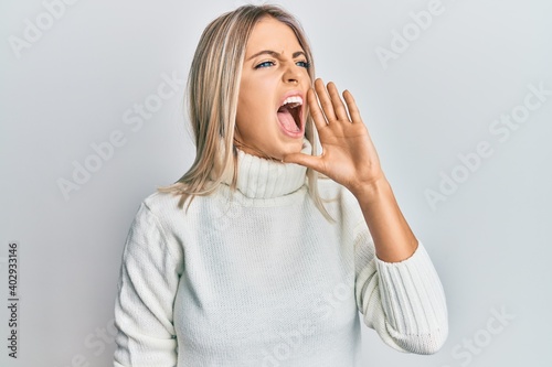Beautiful blonde woman wearing casual turtleneck sweater shouting and screaming loud to side with hand on mouth. communication concept.