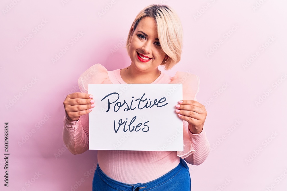Blonde plus size woman asking for optimist attitude holding paper with positive vibes message looking positive and happy standing and smiling with a confident smile showing teeth Blonde plus size woman asking for optimist attitude holding paper with positive vibes message looking positive and happy standing and smiling with a confident smile showing teeth