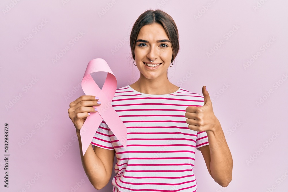 Young hispanic woman holding pink cancer ribbon smiling happy and positive, thumb up doing excellent and approval sign Young hispanic woman holding pink cancer ribbon smiling happy and positive, thumb up doing excellent and approval sign