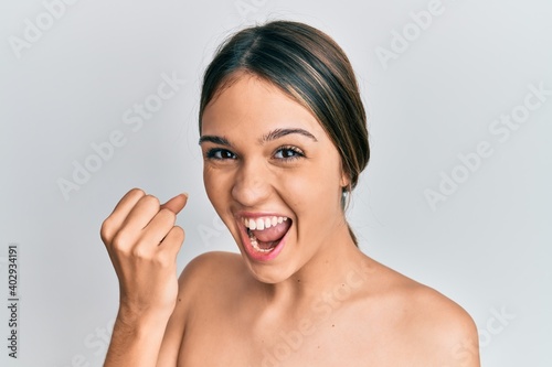 Young brunette woman standing topless showing skin screaming proud, celebrating victory and success very excited with raised arms