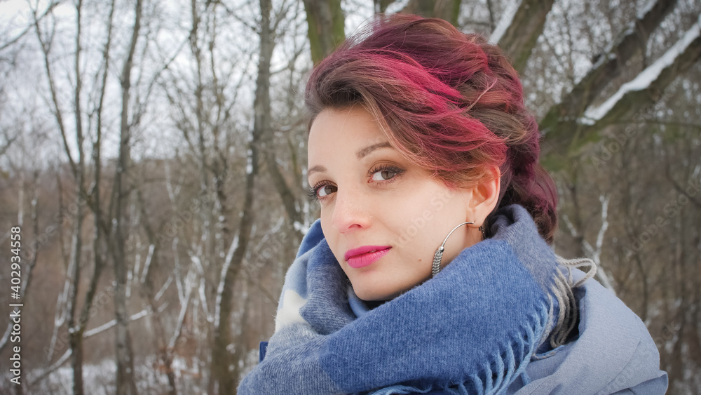 Attractive young woman with perfect skin and makeup with dark pink hair is possing on winter park background wearing blue scarf and silver earrings Attractive young woman with perfect skin and makeup with dark pink hair is possing on winter park background wearing blue scarf and silver earrings