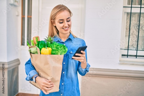 Young blonde girl smiling happy using smartphone and holding grocries paper bag at the city.