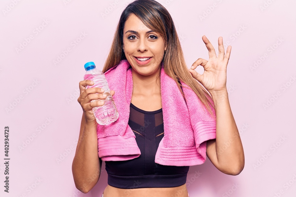 Young beautiful woman wearing sportswear and towel holding bottle of water doing ok sign with fingers, smiling friendly gesturing excellent symbol Young beautiful woman wearing sportswear and towel holding bottle of water doing ok sign with fingers, smiling friendly gesturing excellent symbol