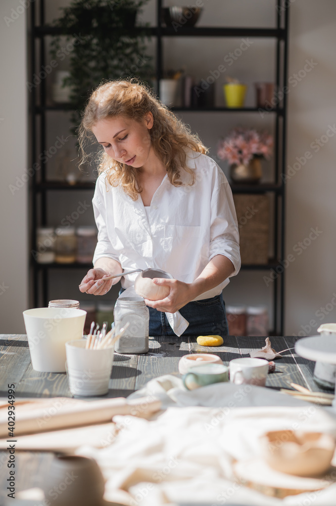 A young european beautiful female potter in a creative process of making ceramics at table in workshop. Hair fluttering in the wind at sunny day at pottery studio. A young european beautiful female potter in a creative process of making ceramics at table in workshop. Hair fluttering in the wind at sunny day at pottery studio.