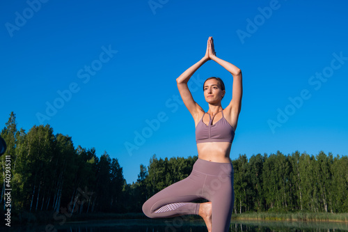 Fototapeta Naklejka Na Ścianę i Meble -  Girl doing yoga tree pose in nature. Girl yoga pose. Sports outdoors stretching on a forest lake.