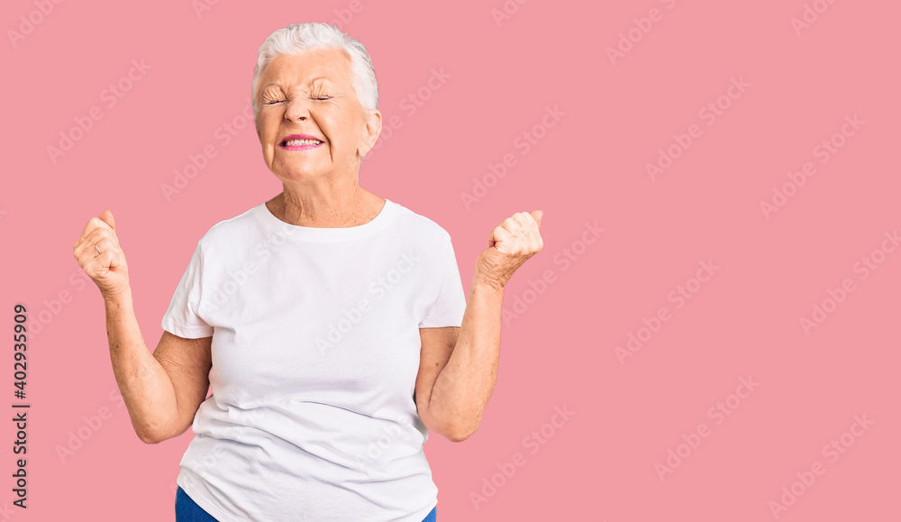 Senior beautiful woman with blue eyes and grey hair wearing casual white tshirt very happy and excited doing winner gesture with arms raised, smiling and screaming for success. celebration concept. Senior beautiful woman with blue eyes and grey hair wearing casual white tshirt very happy and excited doing winner gesture with arms raised, smiling and screaming for success. celebration concept.