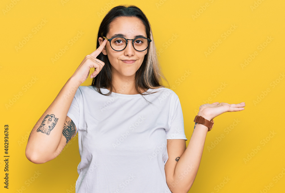 Young hispanic woman wearing casual white t shirt confused and annoyed with open palm showing copy space and pointing finger to forehead. think about it. Young hispanic woman wearing casual white t shirt confused and annoyed with open palm showing copy space and pointing finger to forehead. think about it.