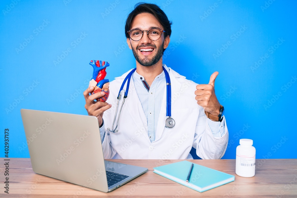 Handsome hispanic man wearing doctor uniform holding heart smiling happy and positive, thumb up doing excellent and approval sign Handsome hispanic man wearing doctor uniform holding heart smiling happy and positive, thumb up doing excellent and approval sign
