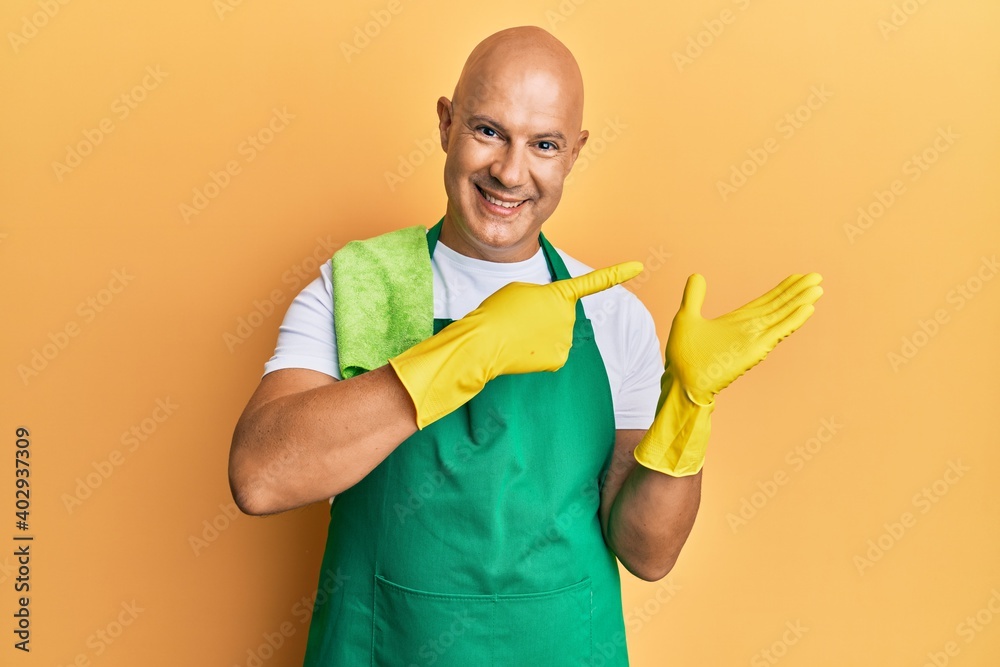 Middle age bald man wearing apron holding cleaning gloves amazed and smiling to the camera while presenting with hand and pointing with finger. Middle age bald man wearing apron holding cleaning gloves amazed and smiling to the camera while presenting with hand and pointing with finger.