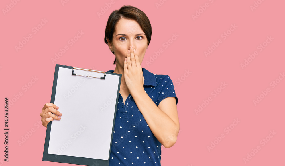 Young brunette woman with short hair holding clipboard with blank space covering mouth with hand, shocked and afraid for mistake. surprised expression Young brunette woman with short hair holding clipboard with blank space covering mouth with hand, shocked and afraid for mistake. surprised expression
