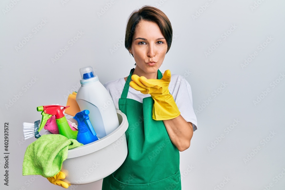 Young brunette woman with short hair wearing apron holding cleaning products looking at the camera blowing a kiss with hand on air being lovely and sexy. love expression. Young brunette woman with short hair wearing apron holding cleaning products looking at the camera blowing a kiss with hand on air being lovely and sexy. love expression.