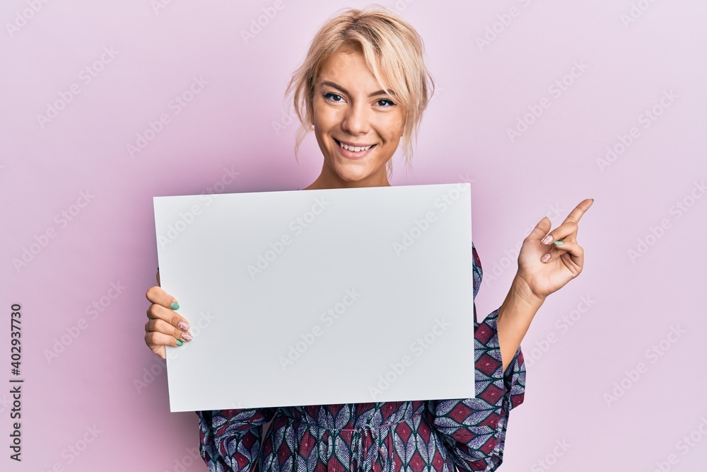 Naklejka premium Young blonde girl holding blank empty banner smiling happy pointing with hand and finger to the side