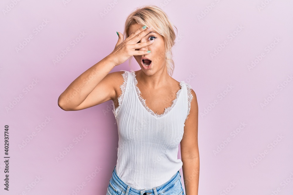 Young blonde girl wearing casual clothes peeking in shock covering face and eyes with hand, looking through fingers with embarrassed expression.