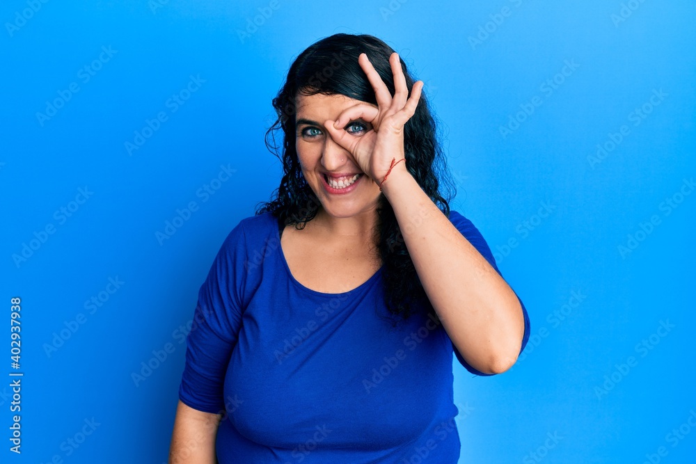 Plus size brunette woman wearing casual blue shirt doing ok gesture with hand smiling, eye looking through fingers with happy face. Plus size brunette woman wearing casual blue shirt doing ok gesture with hand smiling, eye looking through fingers with happy face.