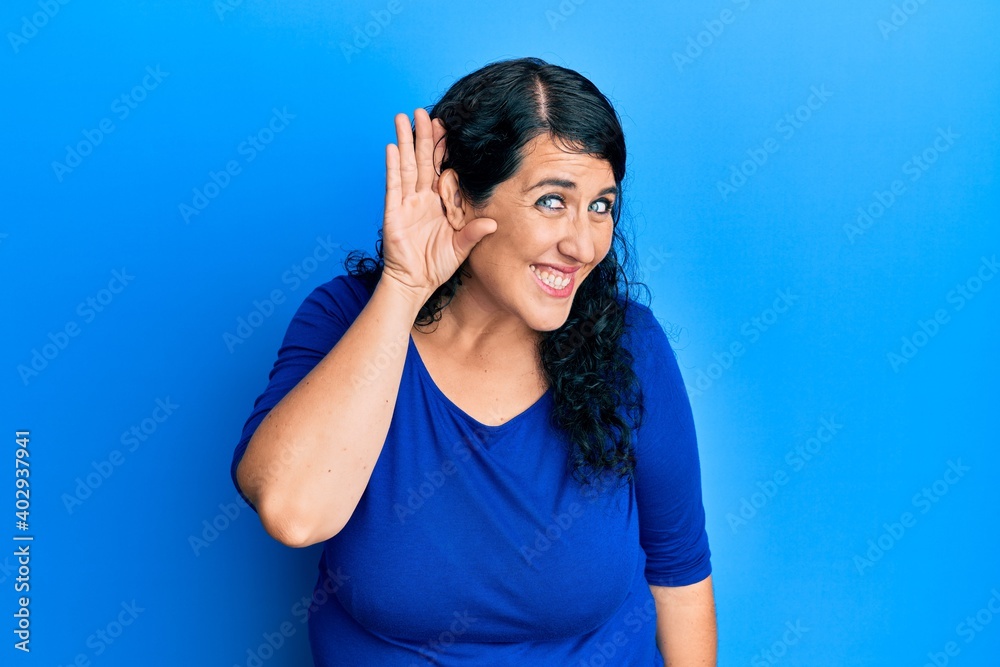 Plus size brunette woman wearing casual blue shirt smiling with hand over ear listening an hearing to rumor or gossip. deafness concept. Plus size brunette woman wearing casual blue shirt smiling with hand over ear listening an hearing to rumor or gossip. deafness concept.