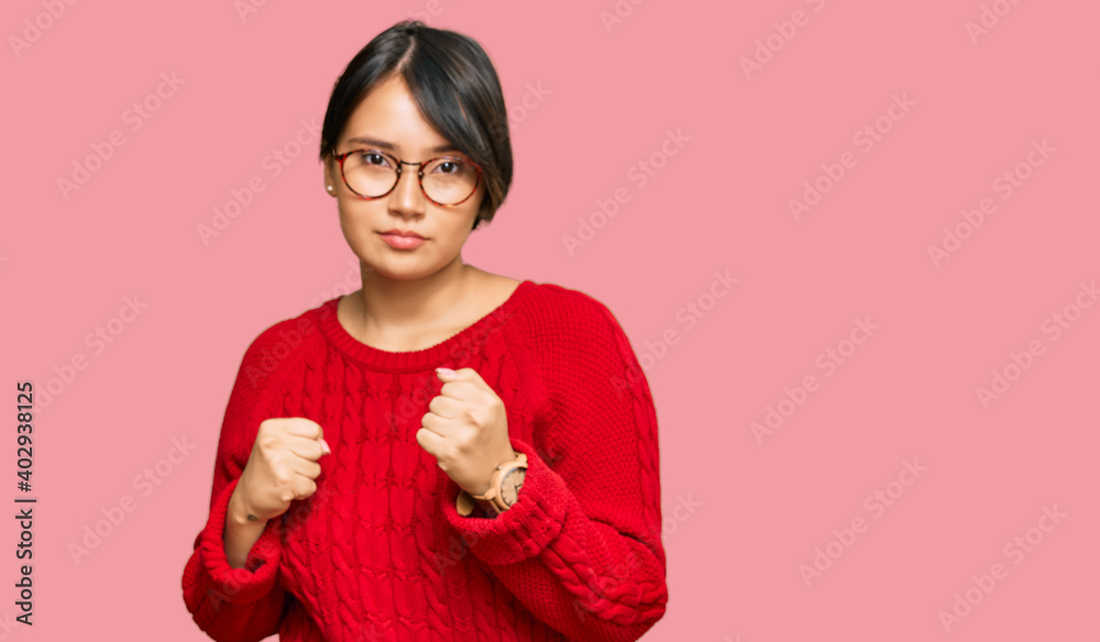 Young beautiful hispanic woman with short hair wearing casual sweater and glasses ready to fight with fist defense gesture, angry and upset face, afraid of problem Young beautiful hispanic woman with short hair wearing casual sweater and glasses ready to fight with fist defense gesture, angry and upset face, afraid of problem