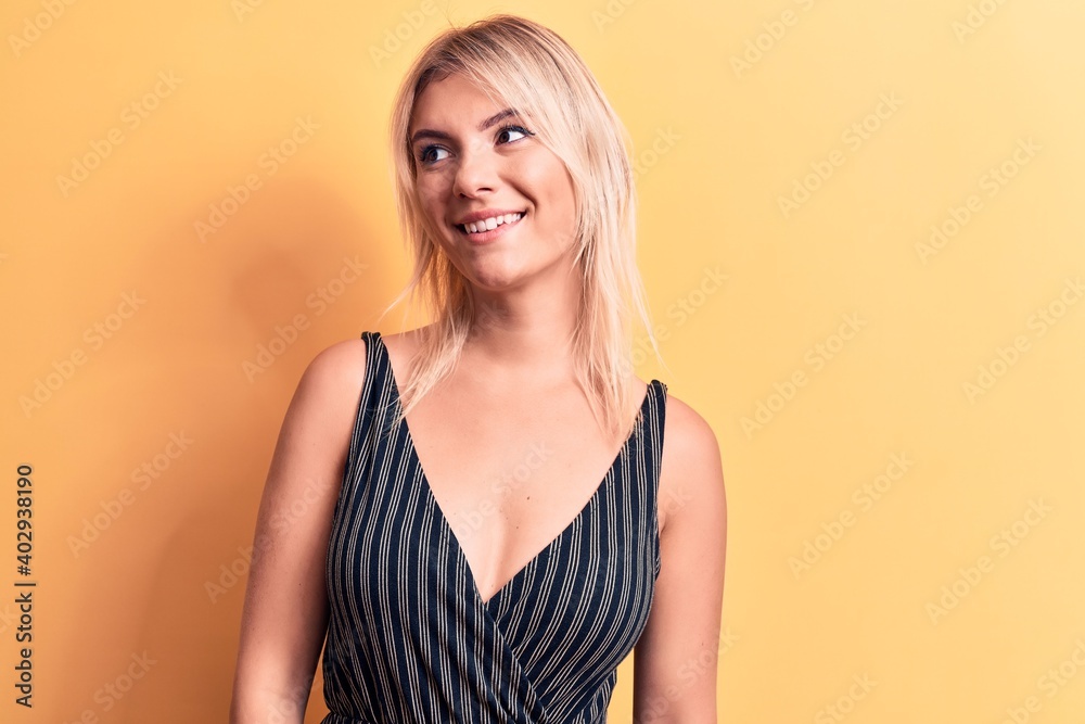 Young beautiful blonde woman wearing striped t-shirt standing over isolated yellow background looking to side, relax profile pose with natural face and confident smile. Young beautiful blonde woman wearing striped t-shirt standing over isolated yellow background looking to side, relax profile pose with natural face and confident smile.