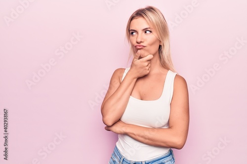 Young beautiful blonde woman wearing casual t-shirt standing over isolated pink background thinking concentrated about doubt with finger on chin and looking up wondering