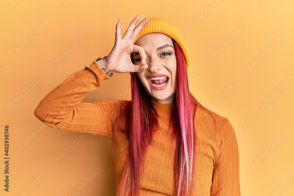 Young caucasian woman wearing wool cap smiling happy doing ok sign with hand on eye looking through fingers Young caucasian woman wearing wool cap smiling happy doing ok sign with hand on eye looking through fingers