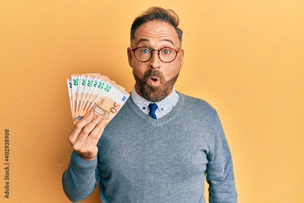 Handsome middle age man holding bunch of 50 euro banknotes scared and amazed with open mouth for surprise, disbelief face - obrazy, fototapety, plakaty 