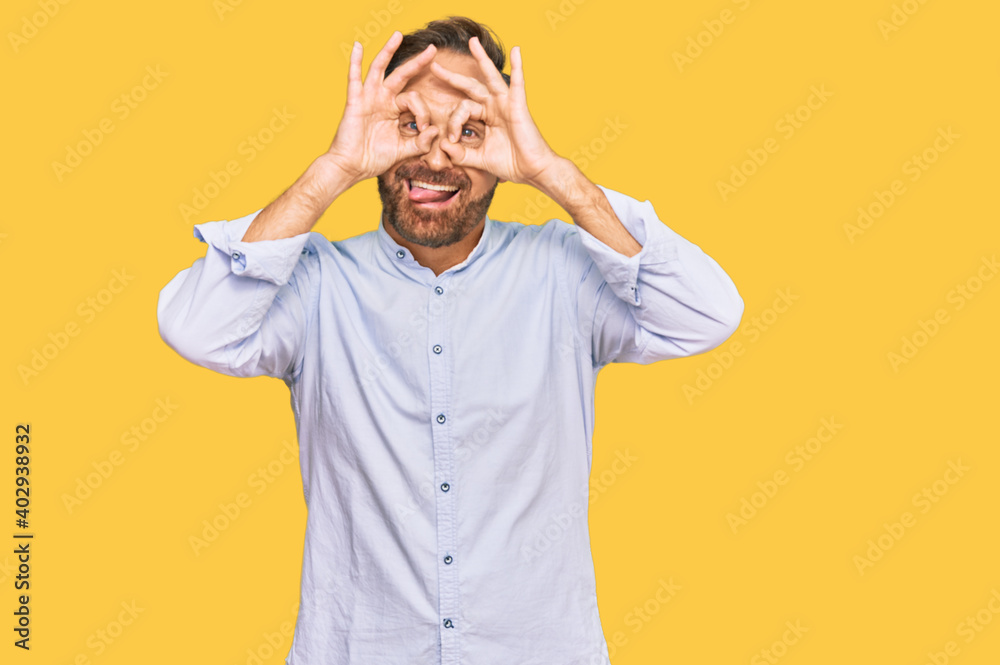 Handsome middle age man wearing business shirt doing ok gesture like binoculars sticking tongue out, eyes looking through fingers. crazy expression.