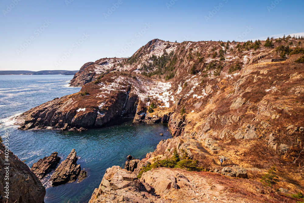 A rugged mountain coastline on a sunny day in Spring.  The cold Atlantic ocean and the sky are deep blues.  The hills and mountains are mostly rocky with some greenery.  The cliffs are a red texture.