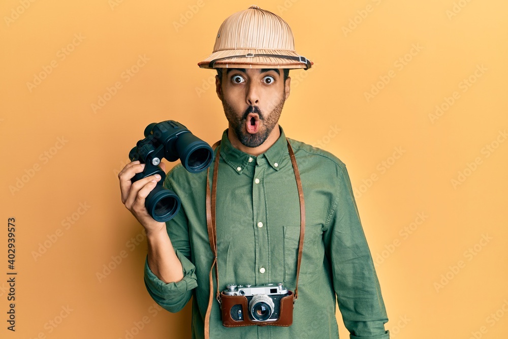 Young hispanic man wearing explorer hat looking through binoculars scared and amazed with open mouth for surprise, disbelief face - obrazy, fototapety, plakaty 