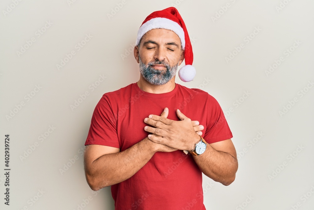 Middle age handsome man wearing christmas hat and summer t-shirt smiling with hands on chest, eyes closed with grateful gesture on face. health concept. Middle age handsome man wearing christmas hat and summer t-shirt smiling with hands on chest, eyes closed with grateful gesture on face. health concept.
