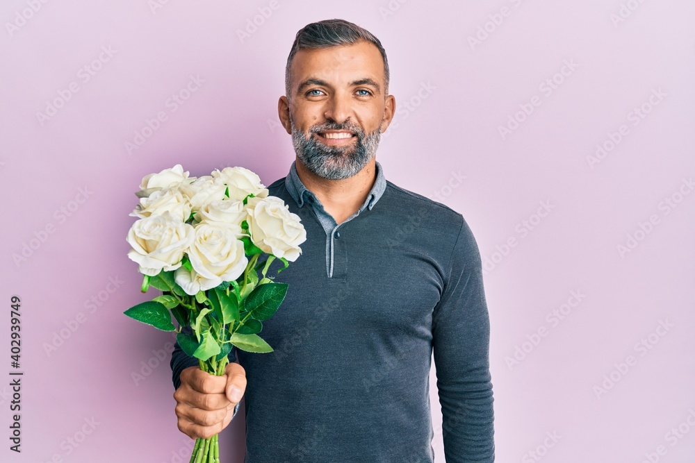 Middle age handsome man holding bouquet of white flowers looking positive and happy standing and smiling with a confident smile showing teeth Middle age handsome man holding bouquet of white flowers looking positive and happy standing and smiling with a confident smile showing teeth