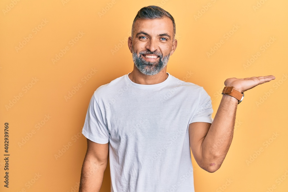 Middle age handsome man wearing casual white tshirt smiling cheerful presenting and pointing with palm of hand looking at the camera. Middle age handsome man wearing casual white tshirt smiling cheerful presenting and pointing with palm of hand looking at the camera.