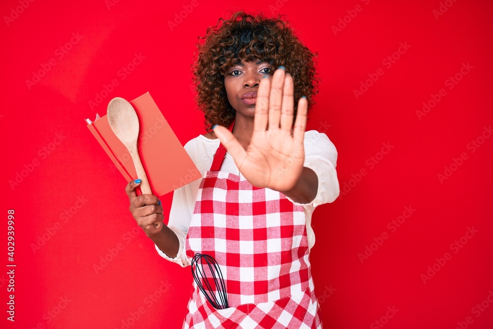 Young african american woman wearing professional baker apron reading cooking recipe book with open hand doing stop sign with serious and confident expression, defense gesture Young african american woman wearing professional baker apron reading cooking recipe book with open hand doing stop sign with serious and confident expression, defense gesture