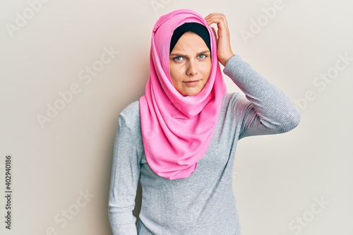 Young caucasian woman wearing traditional islamic hijab scarf confuse and wonder about question. uncertain with doubt, thinking with hand on head. pensive concept.