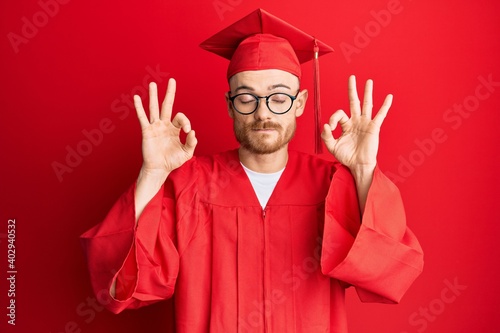 Young redhead man wearing red graduation cap and ceremony robe relax and smiling with eyes closed doing meditation gesture with fingers. yoga concept.