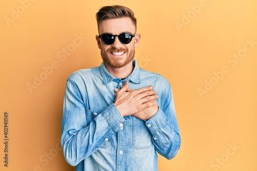 Young redhead man wearing stylish sunglasses smiling with hands on chest with closed eyes and grateful gesture on face. health concept.