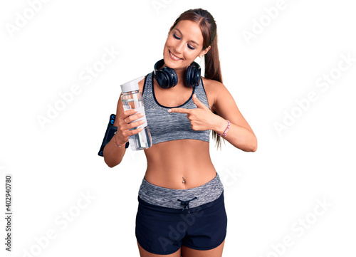 Young beautiful hispanic woman wearing sportswear holding water bottle smiling happy pointing with hand and finger