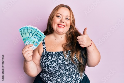 Plus size caucasian young woman holding 50 yuan chinese banknotes smiling happy and positive, thumb up doing excellent and approval sign photo