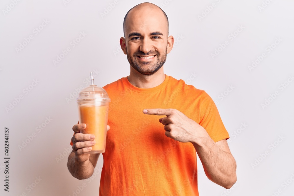 Young handsome bald man drinking glass of healthy orange juice smiling happy pointing with hand and finger Young handsome bald man drinking glass of healthy orange juice smiling happy pointing with hand and finger
