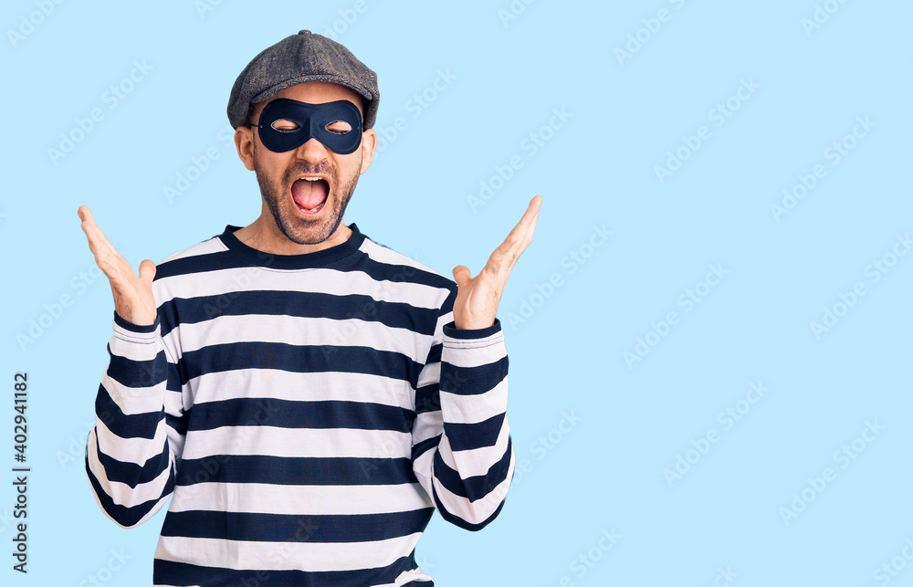 Young handsome man wearing burglar mask celebrating mad and crazy for success with arms raised and closed eyes screaming excited. winner concept Young handsome man wearing burglar mask celebrating mad and crazy for success with arms raised and closed eyes screaming excited. winner concept