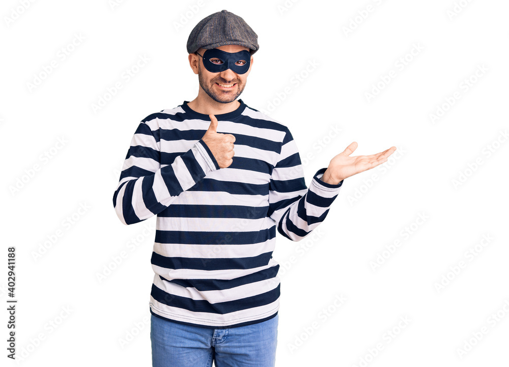 Fototapeta premium Young handsome man wearing burglar mask showing palm hand and doing ok gesture with thumbs up, smiling happy and cheerful