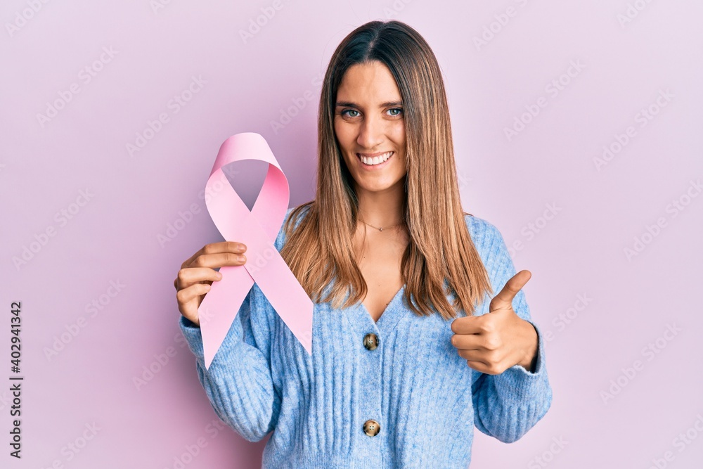 Brunette young woman holding pink cancer ribbon smiling happy and positive, thumb up doing excellent and approval sign Brunette young woman holding pink cancer ribbon smiling happy and positive, thumb up doing excellent and approval sign