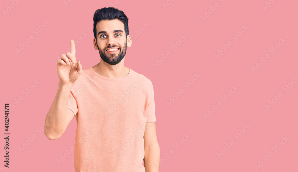Young handsome man with beard wearing casual t-shirt showing and pointing up with finger number one while smiling confident and happy. Young handsome man with beard wearing casual t-shirt showing and pointing up with finger number one while smiling confident and happy.